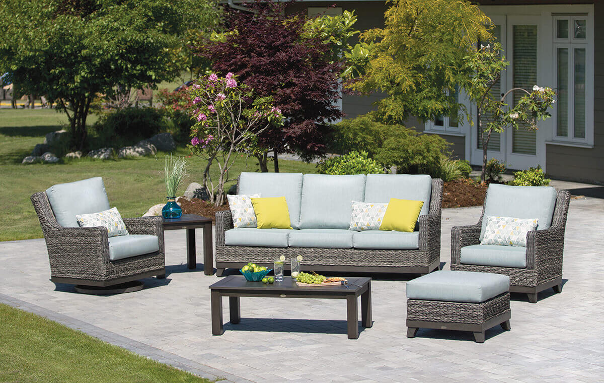 Outdoor patio scene featuring the Asheville Furniture Collection arranged on a grey stone patio with a lush garden and house in the background. The set includes a high-back three-seat sofa with grey Sunbrella cushions, two matching deep seating lounge chairs, a woven resin wicker ottoman, a rectangular dark-brown coffee table, and a square side table with a decorative vase. Bright accent pillows in yellow and geometric patterns add contrast, creating a modern coastal look.