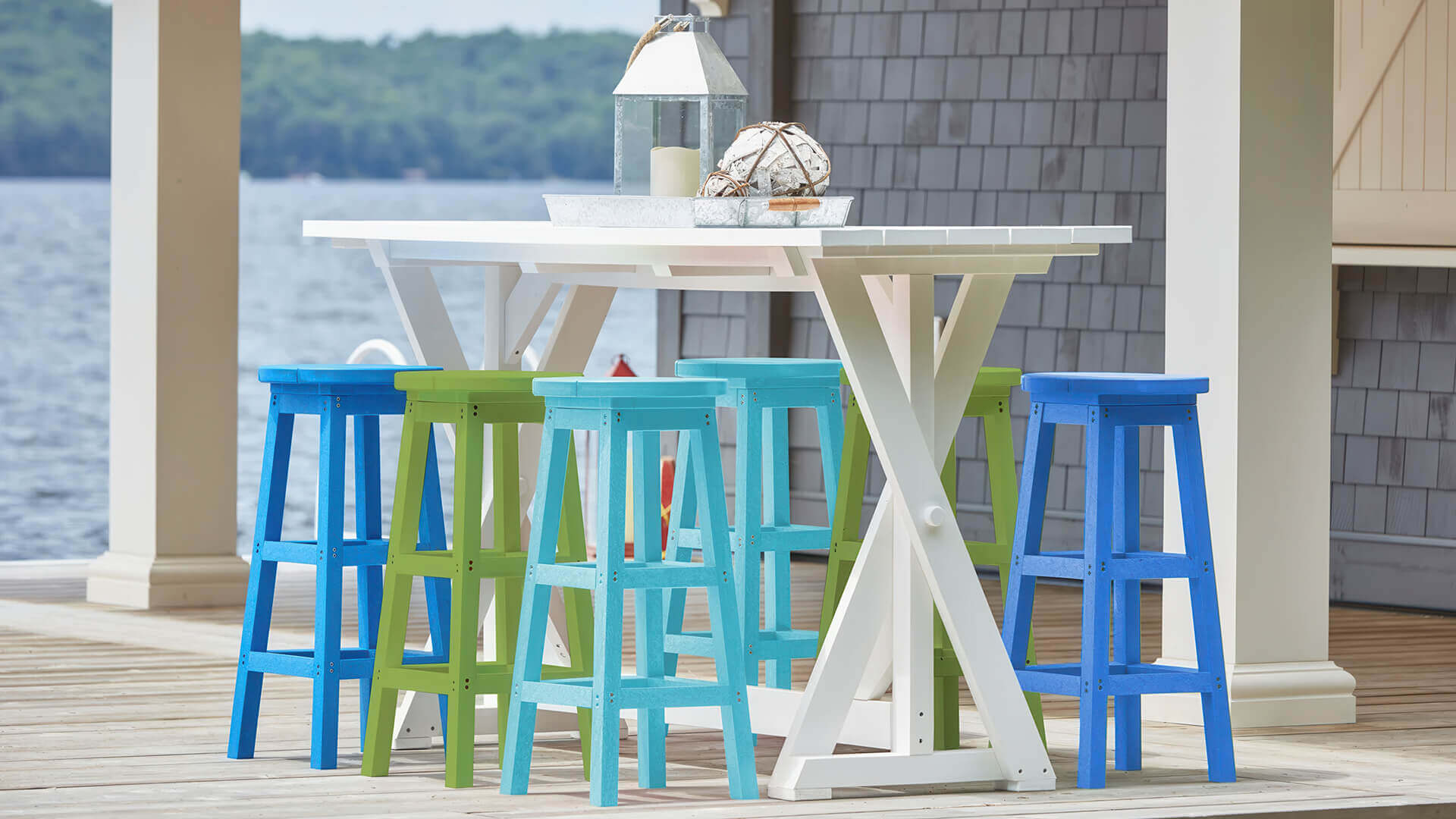 Outdoor lakefront dining setup featuring a white CR Plastics Harvest Bar Table with a trestle-style base and wide plank tabletop, surrounded by six CRP Backless Bar Stools from the CRP Bar Collection. The stools are shown in a mix of bright designer colours, including lime green, turquoise blue, and cobalt blue, contrasting against the crisp white table. The bar-height furniture is placed on a wooden deck overlooking calm lake water, with shoreline trees blurred in the background. Decorative accents on the table include a glass lantern with a large candle and a nautical-style rope-wrapped glass float, enhancing the coastal outdoor aesthetic. The stools’ square, reinforced legs with footrests are visible beneath the table, highlighting their sturdy, commercial-grade design for casual dining and entertaining.