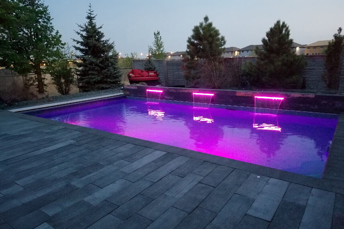 This evening photograph captures a luxurious Winnipeg backyard showcasing the modern Cypress Ridge custom pool with LED waterfalls. The geometric rectangular swimming pool occupies the centre of the scene, surrounded by a premium dark stone deck made of large, staggered rectangular pavers that fill the foreground and side edges. A white integrated cover track system runs straight along the left inside edge of the pool coping. At the far end of the water, a raised feature wall built from dark grey textured stone displays three sheer descent water features. These cascading waterfalls are brilliantly illuminated with bright pink and purple LED lights, creating a vibrant, glowing reflection across the entire surface of the dark pool water. Just behind the left side of the stone feature wall, a large round outdoor lounge bed covered in bright red cushions provides a dedicated seating area. The background environment consists of a dark wooden privacy fence on the right and a metal chain link fence on the left, interspersed with tall green coniferous pine trees and leafy deciduous trees. Beyond the fence line, the dark silhouettes of multiple suburban residential houses are visible against a soft, dusky grey twilight sky.