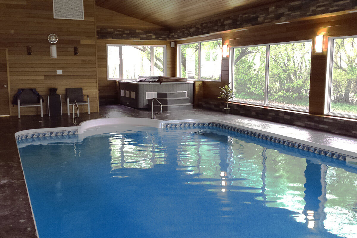 This high-angle interior photograph showcases a luxurious Dacotah custom indoor pool with hot tub, housed within a climate-controlled room featuring warm wood-clad walls and stone-accented bases. The bright blue swimming pool features a crisp patterned liner and a smooth white moulded entry step area positioned in the foreground. In the background, a custom-built Bali tub from Crown Spas & Pools sits on a raised platform, covered by a dark grey thermal lid and accessed by a three-step staircase. The surrounding deck is finished with a dark, textured material that provides a durable surface for the room. Large windows run along the right and rear walls, allowing natural light to illuminate the space while offering views of the exterior landscape. Two chairs and a small table are arranged against the left wall, and a single decorative potted plant stands near the corner of the hot tub platform to complete the wellness oasis.