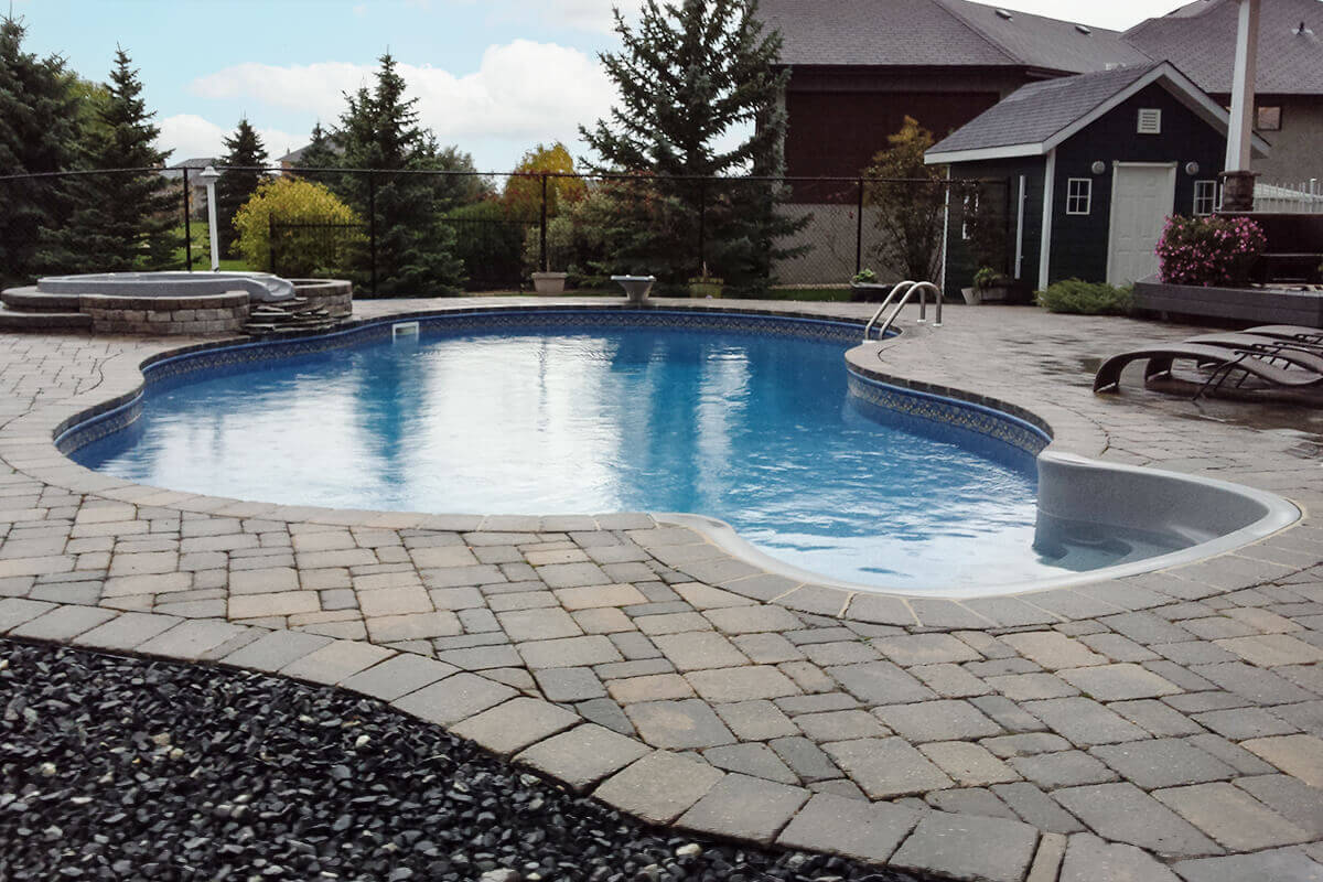 This wide angle photograph showcases a complete backyard transformation featuring a beautiful Doral custom freeform pool with spa. The bright blue swimming pool dominates the centre of the image and is framed by a massive interlocking block patio in shades of grey and tan. On the left side, an elevated grey hot tub structural element is built into a custom stone retaining wall, with stacked natural rock slabs creating a cascading waterfall that flows directly into the main swimming area. The bottom left corner features a distinct border of dark decorative rock beds adjacent to the patio stones. On the right side, curved grey pool steps lead into the water near a stainless steel pool ladder with two handles, alongside several dark brown lounge chairs resting on the wet patio. A sleek black safety fence encloses the area, separating the stone patio from the background environment which includes tall evergreen trees, a dark green residential storage shed with white trim, a large house, and a partly cloudy blue sky.