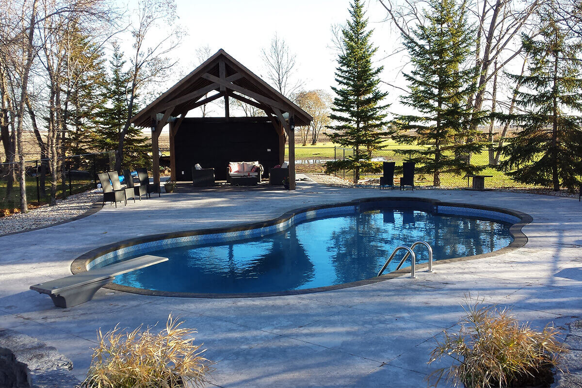 This wide angle outdoor photograph showcases the stunning Waverley custom freeform pool with pavilion set within an expansive backyard layout. The freeform swimming pool occupies the centre right area, filled with clear blue water that features a dark contrasting border and a blue mosaic tile waterline. A grey diving board is installed on the lower left concrete deck, extending over the deep end of the water, while a stainless steel ladder with two curved handrails sits on the opposite right side. A massive textured concrete deck in a light grey finish covers the entire foreground and surrounds the pool, accented by two large potted ornamental grasses at the very bottom edge. In the upper left background, a large open heavy-timber wooden pavilion stands adjacent to the pool, housing a comfortable outdoor lounge area with dark wicker seating and light cushions. The surrounding environment consists of a manicured green lawn, tall evergreen pine trees to the right, bare deciduous trees to the left, and a distant open field under a bright clear sky.