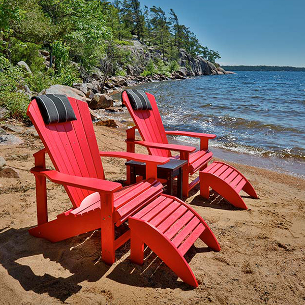 Pair of Classic Adirondack Chairs from Crown Spas & Pools’ Adirondack Furniture Collection shown in bold red finish positioned directly on a sandy beach shoreline. Each chair is equipped with a matching red Standard Foot Stool that extends outward with curved slats for leg support. Both chairs have tall slatted backs and wide contoured seats, complemented by detachable black headrest cushions placed at the top of the backrests. A small square black side table sits between the chairs for convenience. The scene captures waves rolling in against rocky shoreline in the background under a bright blue sky, highlighting the chairs as both stylish and practical for waterfront relaxation.