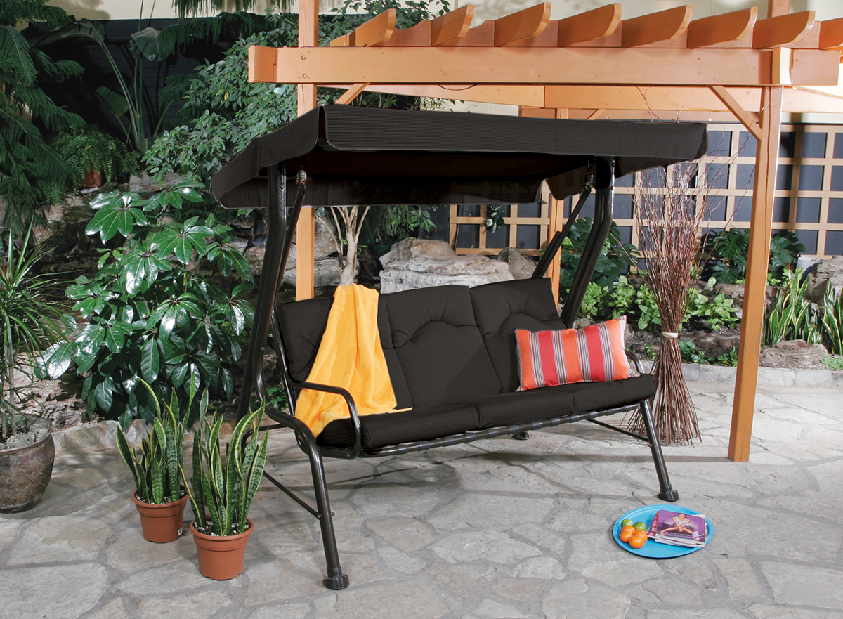 Outdoor three-seat steel swing with a black cushioned seat and backrest under a matching black water-resistant canopy. The frame is powder-coated steel with curved armrests, set on a stone patio beneath a wooden pergola. A yellow towel is draped over the left seat, and a striped orange-and-grey accent cushion rests on the right side. Decorative plants, branches, and a blue tray with oranges and a magazine enhance the relaxing setting, making the swing ideal for backyard leisure and shade.