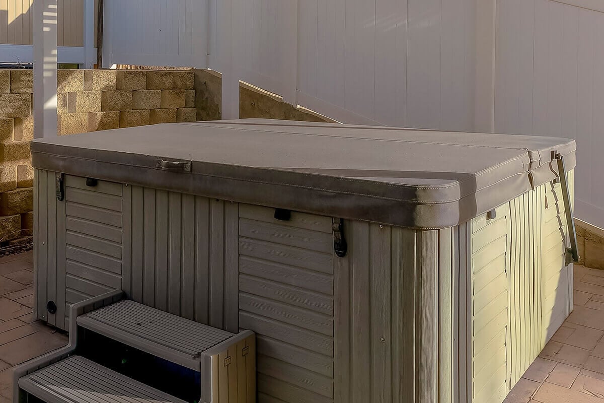 Light grey marine grade vinyl hot tub cover fitted on an outdoor spa with steps and side lifter hardware.