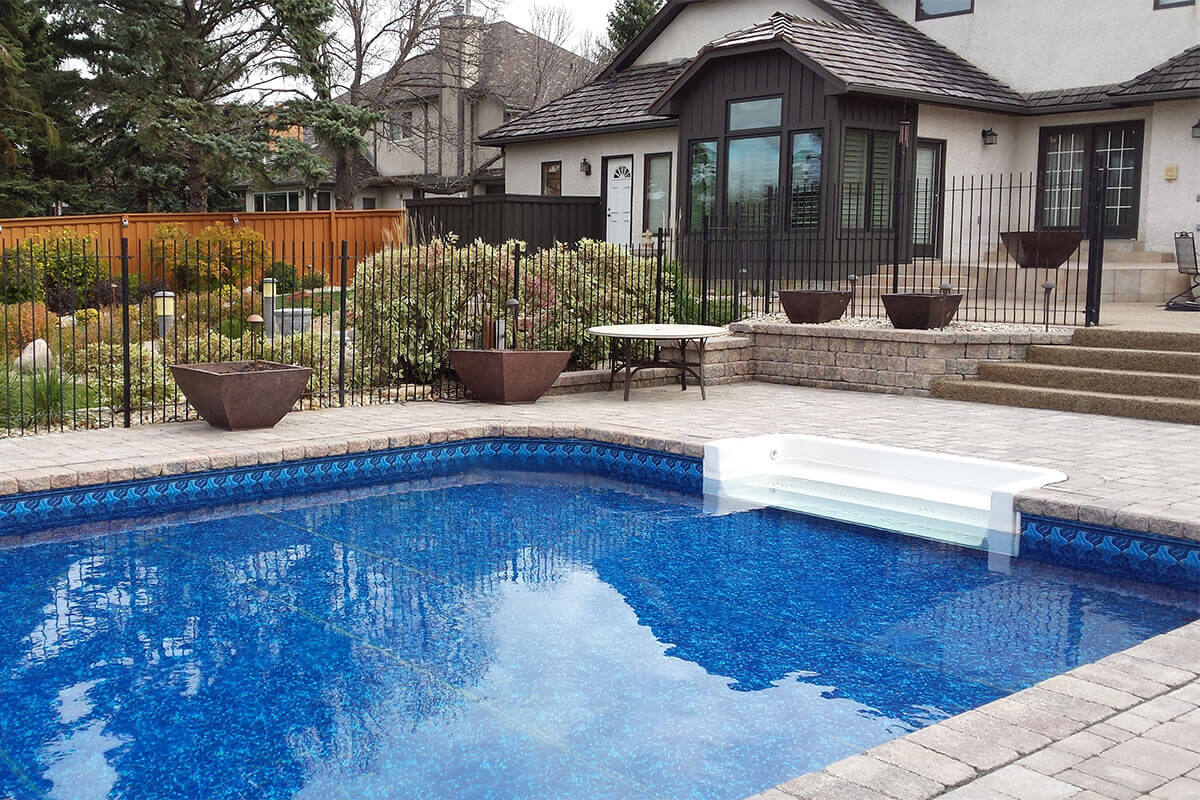 This angled outdoor photograph showcases the shallow end of the luxurious Shorecrest inground pool with paving stones. The bright blue swimming pool water fills the bottom left, featuring a distinct patterned blue liner border along the inside edge. On the right side of the water, a crisp white walk-in step section provides effortless entry from the massive, beautifully patterned stone paver deck that surrounds the pool. In the midground, the patterned stone deck transitions into wide concrete steps leading to an upper terrace tier, separated by a low stone retaining wall decorated with several square brown planter bowls. Behind a black metal safety fence, lush green landscaping and a solid brown wooden fence enclose the left side of the yard. On the right, the large beige stucco house features dark brown window trim, multiple doors, and a wood shingle roof under a bright overcast sky.