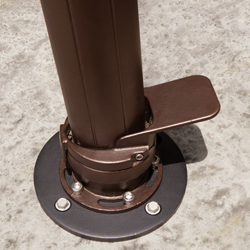 Close-up view of the AKZ Base Concrete Mount Kit, securely fastened onto a textured concrete surface. The circular black metal base plate is bolted down with four silver anchor bolts. A sturdy bronze-coloured umbrella pole rises vertically from the mount, attached through a precision-engineered collar featuring a flat locking pedal mechanism for stability. The metallic textures, tight bolts, and clean edges emphasise the robust construction of the mount, designed to keep patio umbrellas firmly locked in place on concrete installations for maximum safety and durability.