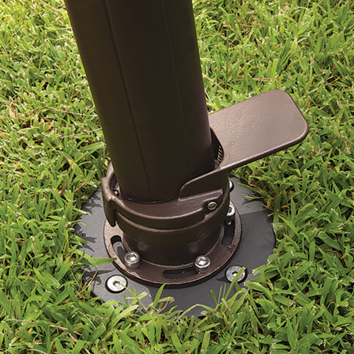 Close-up view of the AKZ In Ground Mount Kit securely installed within a lush green lawn. The dark bronze umbrella pole is fastened to a round black metal base plate, which is anchored flush to the ground with four visible bolts and reinforced slots. The integrated foot pedal mechanism and precision hardware highlight its durable engineering. The surrounding grass slightly overlaps the metal base, showing how the mount blends neatly into outdoor landscapes. This image captures the mount’s purpose of providing a permanent, stable foundation for AKZ or AG25 cantilever umbrellas through in-ground installation within concrete for long-term reliability and strength.