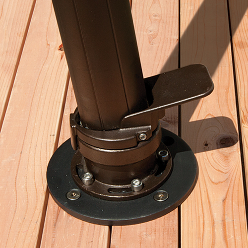 Close-up image of the AKZ Base Wood Mount Kit securely installed on a light natural wood deck surface. The dark bronze umbrella support pole is firmly attached to a circular black steel mounting plate that is bolted into the wooden planks with four visible silver screws. The mount’s integrated foot pedal protrudes slightly from the side of the bracket, designed for effortless locking and release of the umbrella post. The precise hardware fit and polished metallic components highlight the mount’s superior stability and clean, integrated appearance, perfectly suited for securely anchoring AKZ or AG25 cantilever umbrellas to wood deck patios.