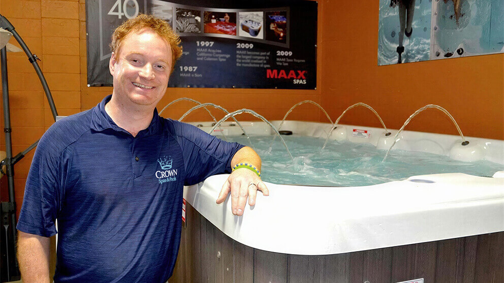 Mike Sudoski Jr. standing beside a filled and operating hot tub at Crown Spas & Pools in Winnipeg. He is positioned on the left side of the image, leaning his right arm casually on the rounded white acrylic edge of the spa, smiling directly toward the camera. He is wearing a navy blue polo shirt embroidered with the Crown Spas & Pools logo on the left chest, reinforcing his role within the family-owned business. To the right of him, the hot tub occupies the foreground, filled with water and actively running, with multiple arcing water features rising from the back edge and curving inward toward the centre of the spa. The interior of the tub is partially visible, showing seating contours beneath the water surface and gentle turbulence from the jets. The cabinet exterior below the rim features a textured wood-style panel in a grey-brown tone. Behind the spa, an orange-toned interior wall displays mounted promotional materials, including a large black timeline-style poster with images and text referencing spa manufacturing history and the MAAX Spas logo in red on the right side. Additional spa imagery appears on the upper right, showing close-up visuals of water and jets. The showroom lighting is warm and even, highlighting both the product and the individual.
