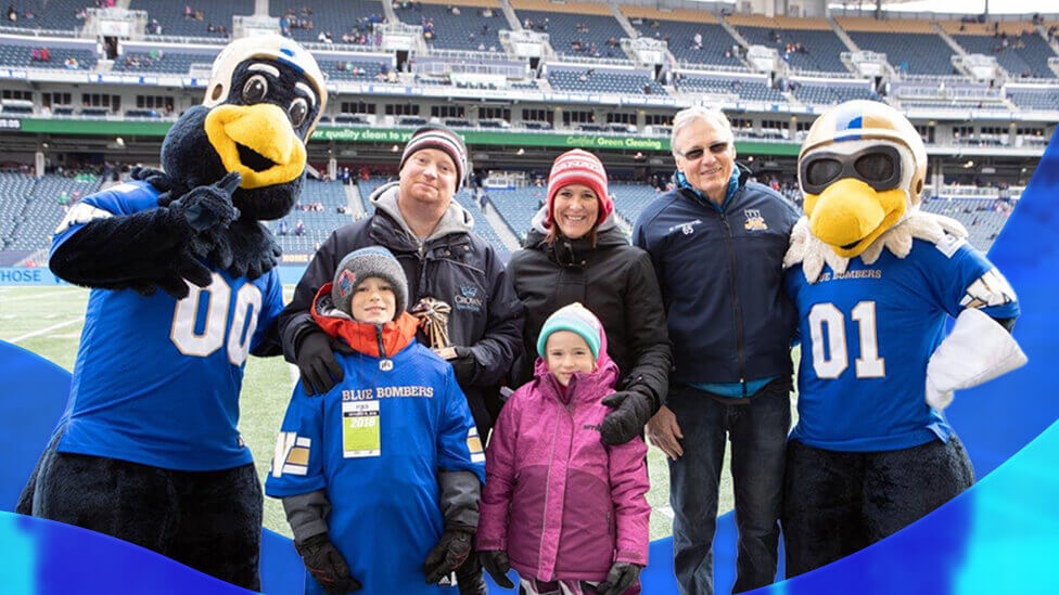 Group photo taken on a football field inside the Princess Auto Stadium in Winnipeg, representing Crown Spas and Pools’ Winnipeg community involvement. Centered in the image is a family of four standing together, bundled in winter clothing, with two adults behind and two children in front. The child on the left wears a blue Winnipeg Blue Bombers jersey layered over winter gear, while the child on the right wears a bright pink winter jacket and patterned snow pants. Flanking the group on both sides are two Winnipeg Blue Bombers bird mascots in full costume, each wearing blue team jerseys with large white numbers, one on the left gesturing toward the camera and one on the right standing upright. Behind the family, two adult men in jackets and hats stand smiling, one holding a trophy. The background shows rows of stadium seating with scattered spectators and structural elements of the arena.