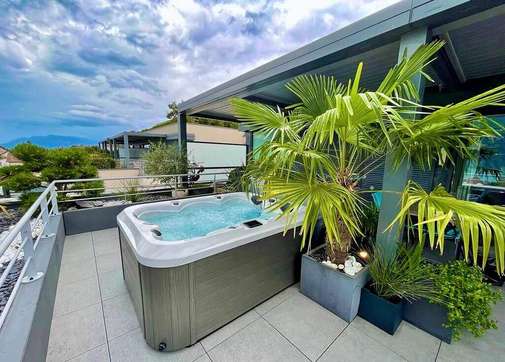 Modern outdoor patio featuring a Maple Spas M320 spa with bubbling water, surrounded by lush tropical plants and sleek contemporary decor, set against a backdrop of mountains and cloudy skies.