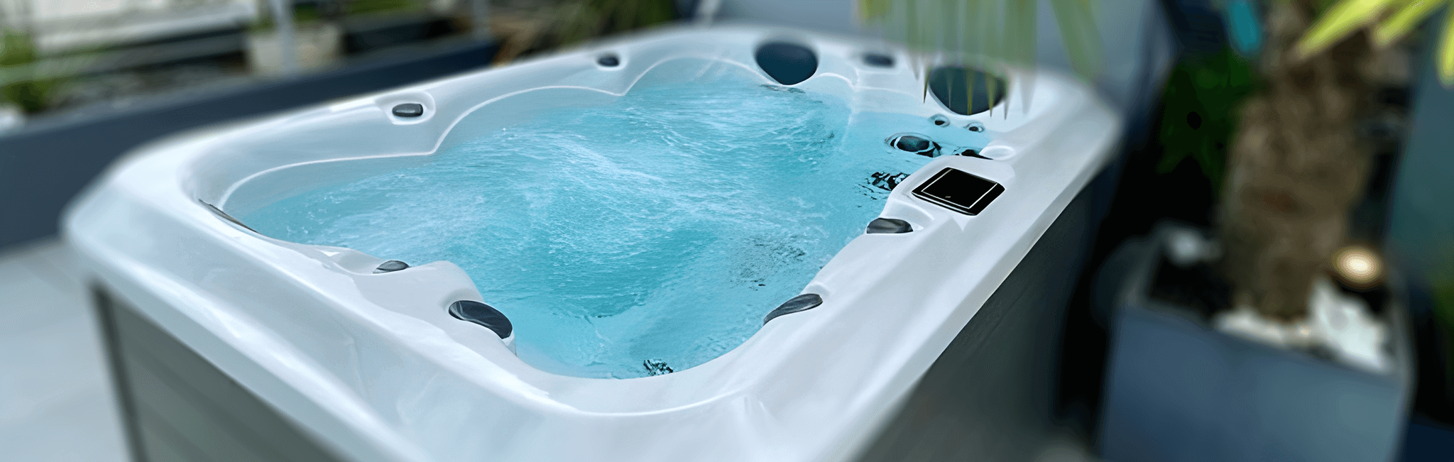 Angled view of a filled hot tub in an outdoor setting, featuring bubbling water, contoured seating with headrests, and built-in jet controls. The modern design includes a sterling acrylic interior and a grey exterior cabinet, with surrounding greenery and patio decor visible in the background.