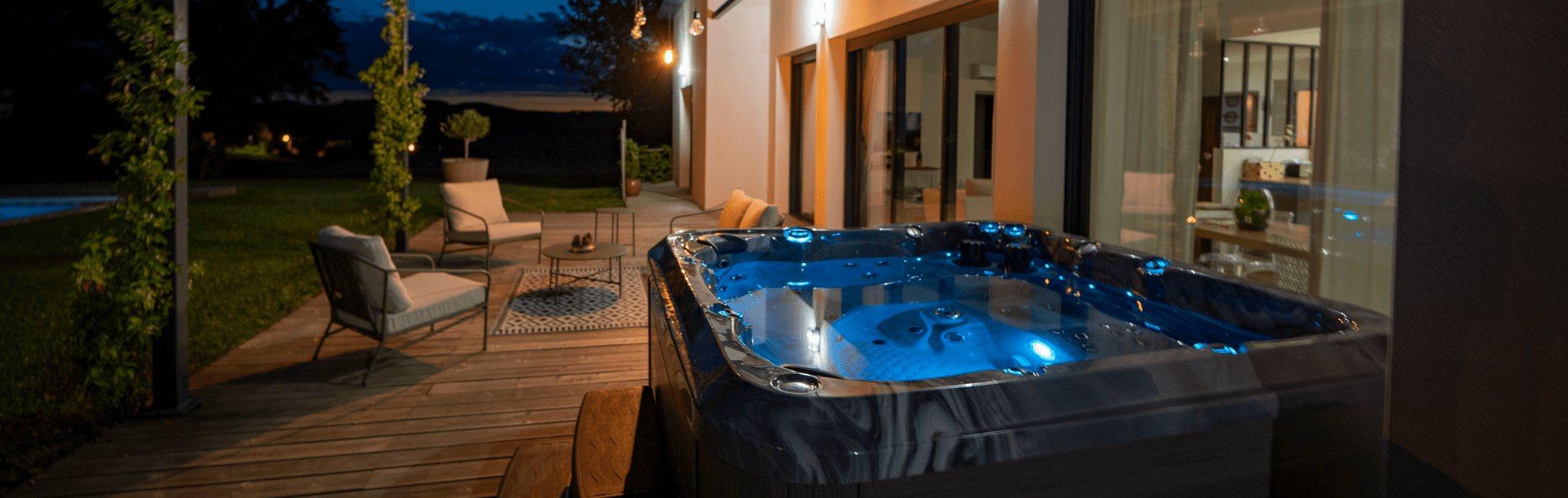 An image of a lighted patio deck at night. On the bottom right of the picture is a hot tub that has a marble pattern and blue lighting underwater and to the left of it are pillars with vines enveloping them. At the back of the lighted patio deck are beige patio chairs with aluminum frames.