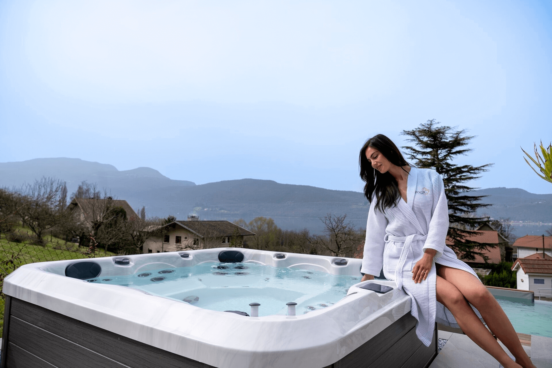 A woman in a white robe sits on the edge of a Maple Spas M420 hot tub, enjoying a serene outdoor setting with mountain views and nearby houses.