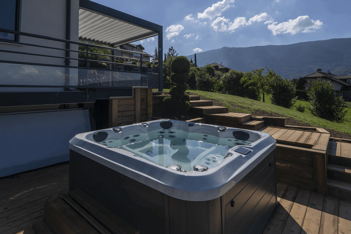 A modern Maple Spas M520 hot tub installed on a wooden deck beside a contemporary home, with built-in seating and multiple jets, set against a scenic backdrop of green hills, trees, and a mountain range under a partly cloudy sky.