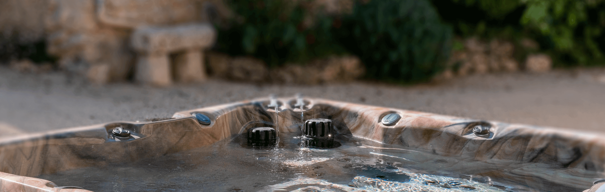 Outdoor view of a Maple Spas M520 hot tub with a marbled brown acrylic shell, gently rippling water, and visible control dials and water jets, set against a rustic stone and garden backdrop for a serene and inviting ambiance.