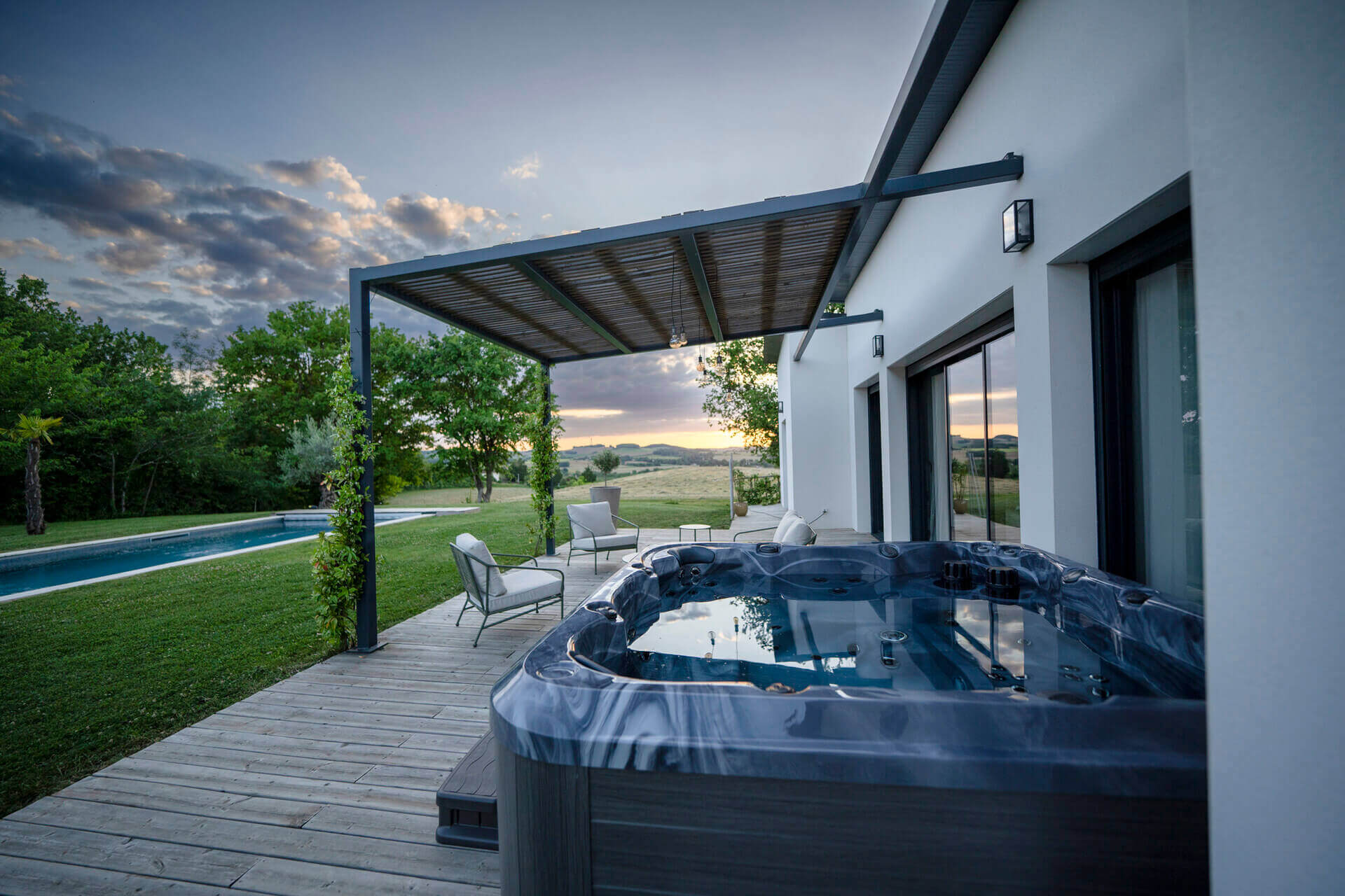 The Maple Spas M620 hot tub setup on a wooden deck next to a contemporary white house, featuring a sleek dark marble-accented spa with water jets, patio seating under a pergola, and scenic views of a grassy yard, trees, and distant hills at sunset.