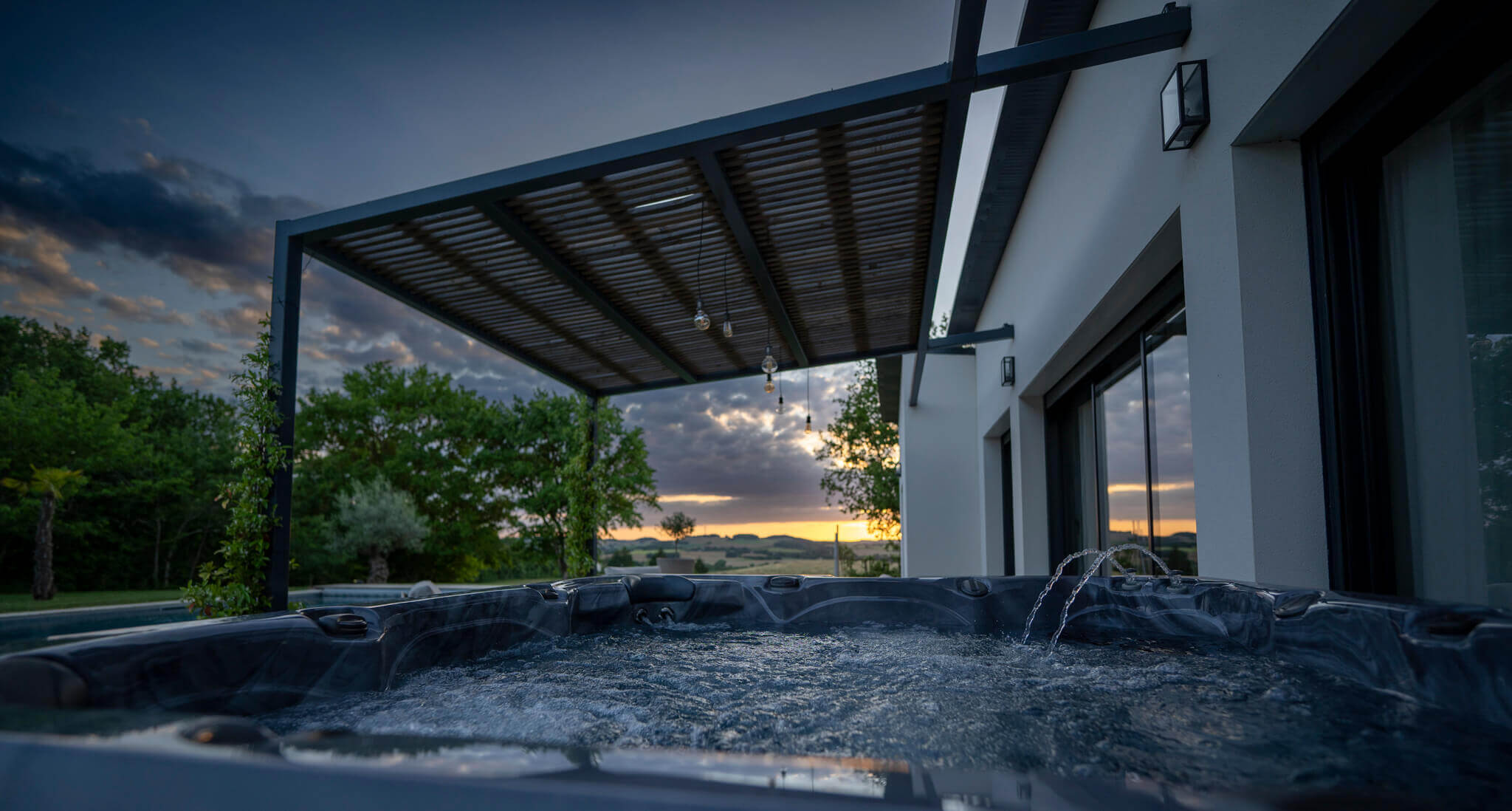 Close-up view of the luxurious Maple Spas M620 hot tub with bubbling water and arcing fountains, set on a deck beside a modern white house, with a pergola overhead and a scenic sunset visible over distant hills and trees in the background.