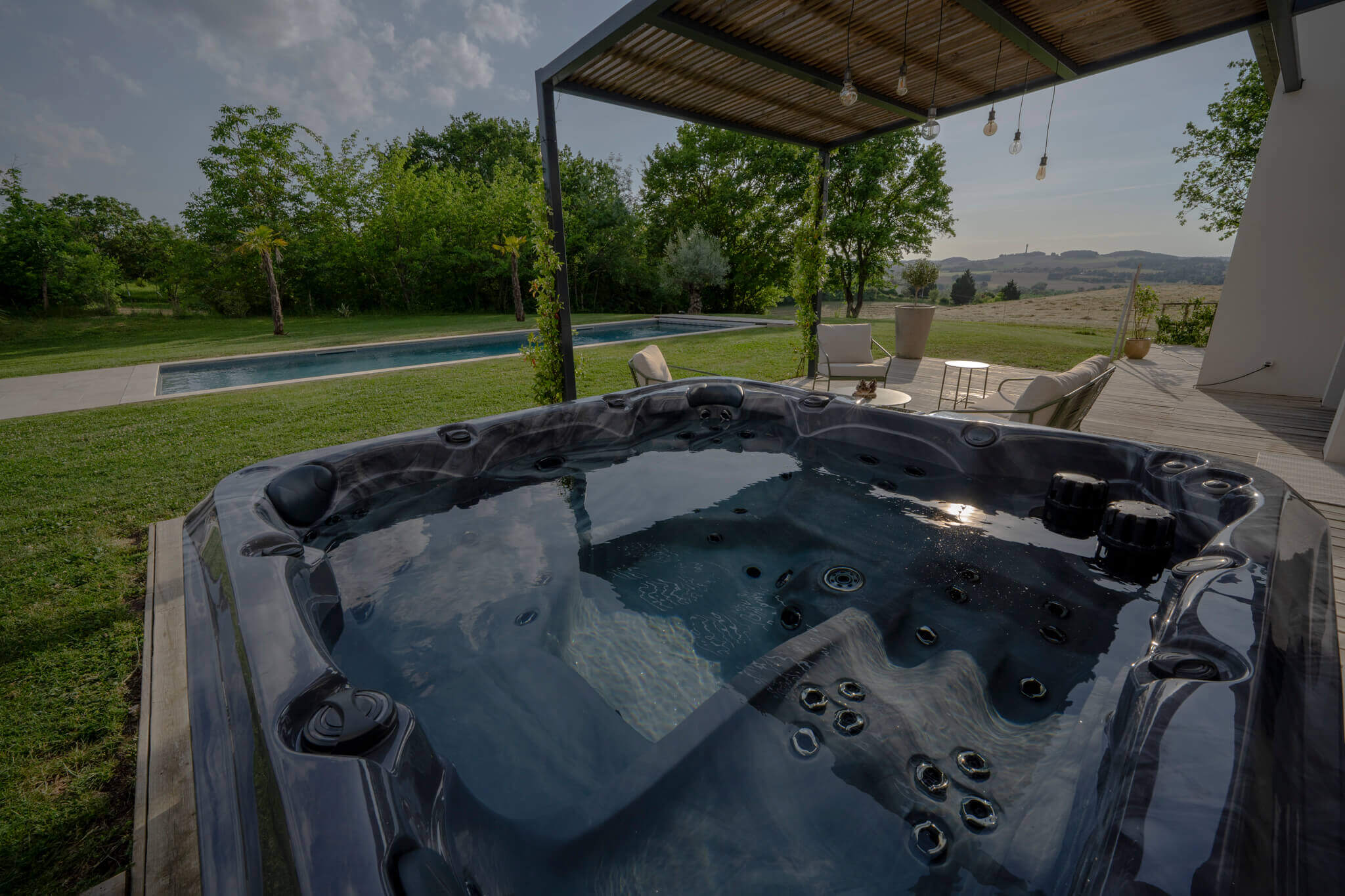 A Maple Spas M620 hot tub positioned on a wooden deck beside a modern home, featuring ergonomic seating and multiple jets, with a shaded pergola above, scenic countryside views in the distance, and a rectangular swimming pool visible on a well-manicured lawn.