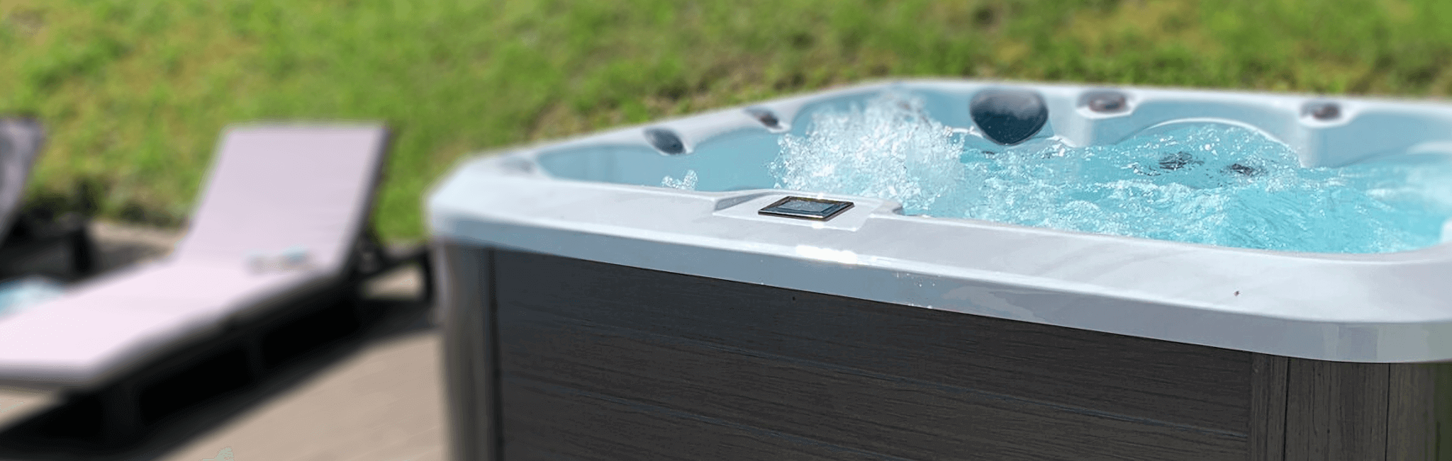 Outdoor view of a Maple Spas M620 hot tub featuring a sleek, modern design with a sterling white acrylic shell, bubbling turquoise water, cushioned headrests, and a digital control panel, set on a wooden deck beside lounge chairs and a grassy landscape.