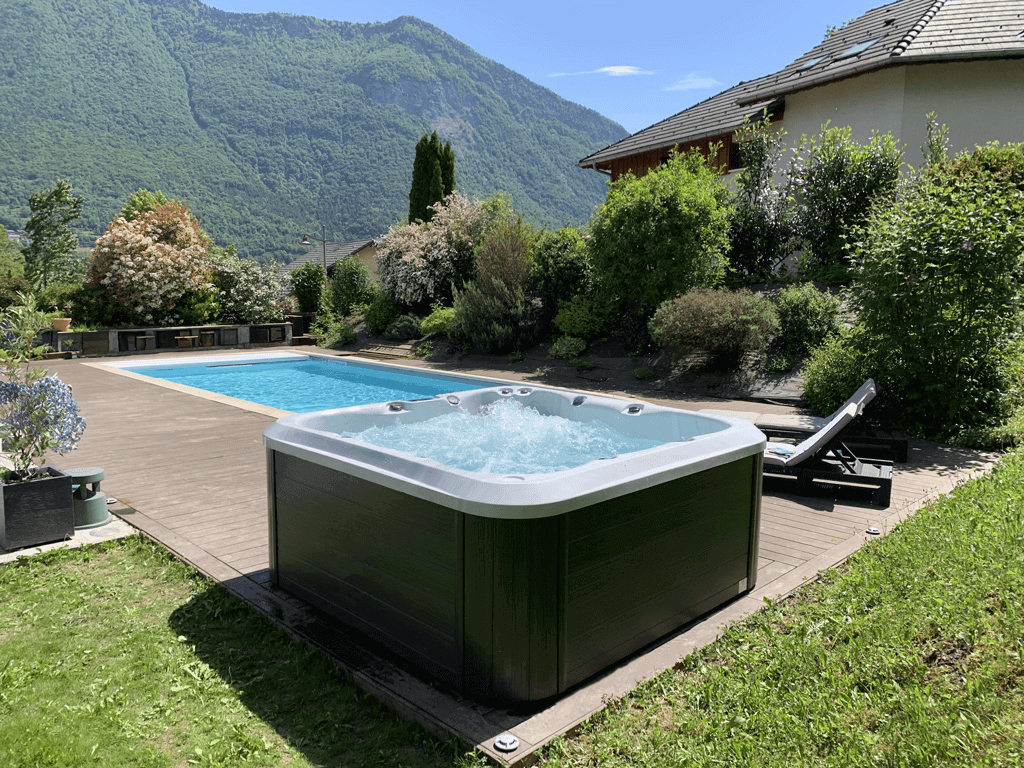 Outdoor view of a bubbling Maple Spas M720 hot tub installed on a wooden deck beside a swimming pool, surrounded by lush greenery, blooming shrubs, and mountain scenery under a clear blue sky.