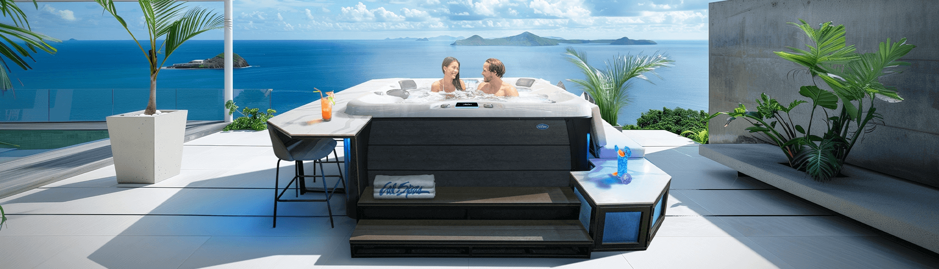 A couple relaxes in a modern Cal Spas hot tub overlooking a stunning ocean view with lush islands in the distance. The hot tub is situated on a sleek, tiled patio with tropical plants, a bar seating area with drinks, and integrated steps. The scene evokes luxury, tranquility, and outdoor comfort, with 'Cal Spas' branding visible on a towel draped over the steps.