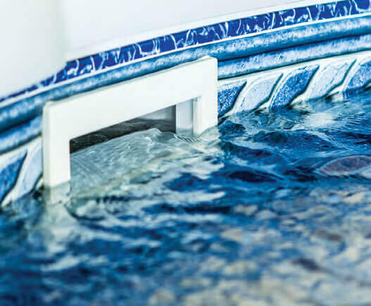 Close-up image of an in-wall skimmer installed on the interior wall of an above ground swimming pool. The rectangular white skimmer opening is partially submerged, with water gently flowing into it. Surrounding the skimmer is a patterned blue vinyl liner featuring a mosaic tile design at the top and a textured wave pattern below. The skimmer helps filter debris from the pool surface by drawing water into the filtration system.