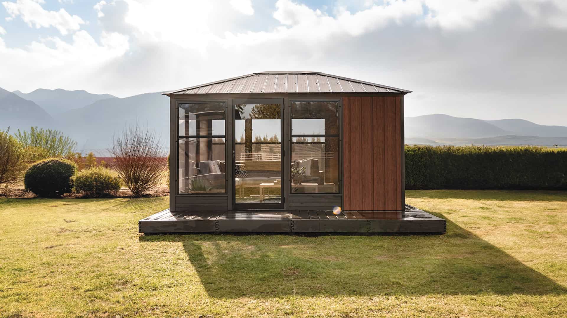Front view of the Adela Gazebo set on a grass lawn, featuring a symmetrical modern design with a black metal 4-sloped roof. The front façade has a central hinged tempered glass door with black aluminium framing, flanked on both sides by tall vinyl-framed windows. The left section of the structure shows floor-to-ceiling glass, while the right side is clad in rich vertical brown wood-style siding. Through the glass, grey cushioned seating with accent pillows and a wooden table are visible inside.