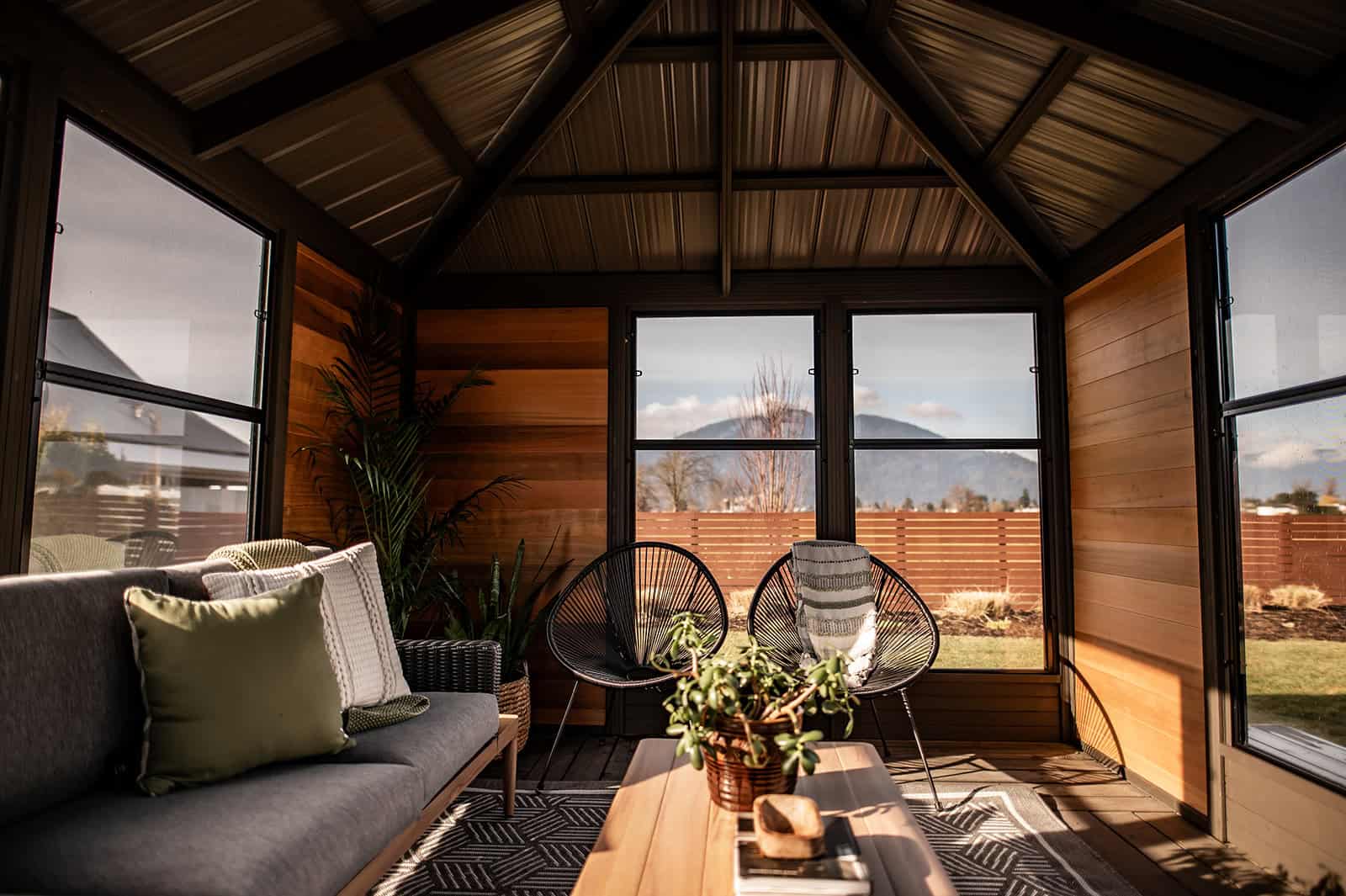Interior view of the Adela Gazebo showcasing a warm and inviting space with natural wood wall cladding and a vaulted black metal roof. On the left side, a modern grey cushioned sofa with green and cream accent pillows sits against the wooden wall. At the far end, two black wire-frame lounge chairs with angled legs face the windows, accompanied by a striped throw blanket draped over one chair. A low rectangular wooden coffee table with a woven plant basket sits in the centre on a geometric patterned grey rug. Tall green plants in wicker baskets are placed in the corners, complementing the natural outdoor view visible through the large tempered glass windows.