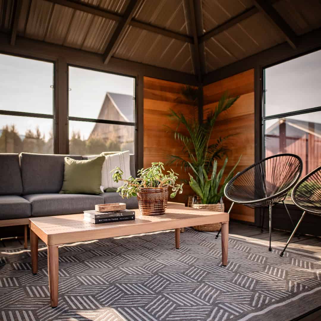 Interior corner view of the Adela Gazebo with warm wooden wall panels and a vaulted black roof. A modern grey sofa with green and cream accent pillows runs along the left wall, positioned behind a rectangular wooden coffee table. The coffee table holds a woven basket with a leafy green plant and stacked books. To the right side, two black round wire-frame chairs with angled legs and woven backs are arranged against the wall. A tall palm-style plant in a wicker basket is tucked into the corner, adding greenery. A geometric grey rug covers the wooden flooring, enhancing the contemporary lounge setting.