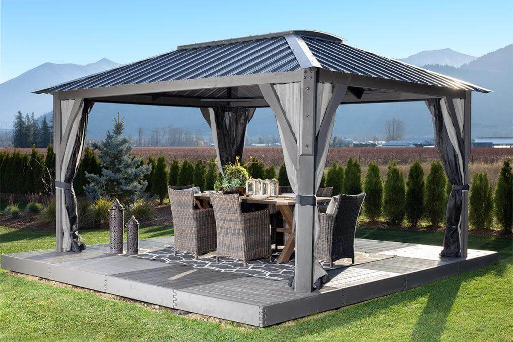 Wide-angle outdoor photograph of the Aluna gazebo installed in a landscaped backyard. The gazebo’s four corner posts rise vertically, supporting a flat rectangular roof with semi-transparent panels that filter sunlight. Beneath the structure sits a full patio lounge set with cushioned chairs and a table, framed by surrounding greenery, illustrating how the Aluna creates a defined, shaded social space.