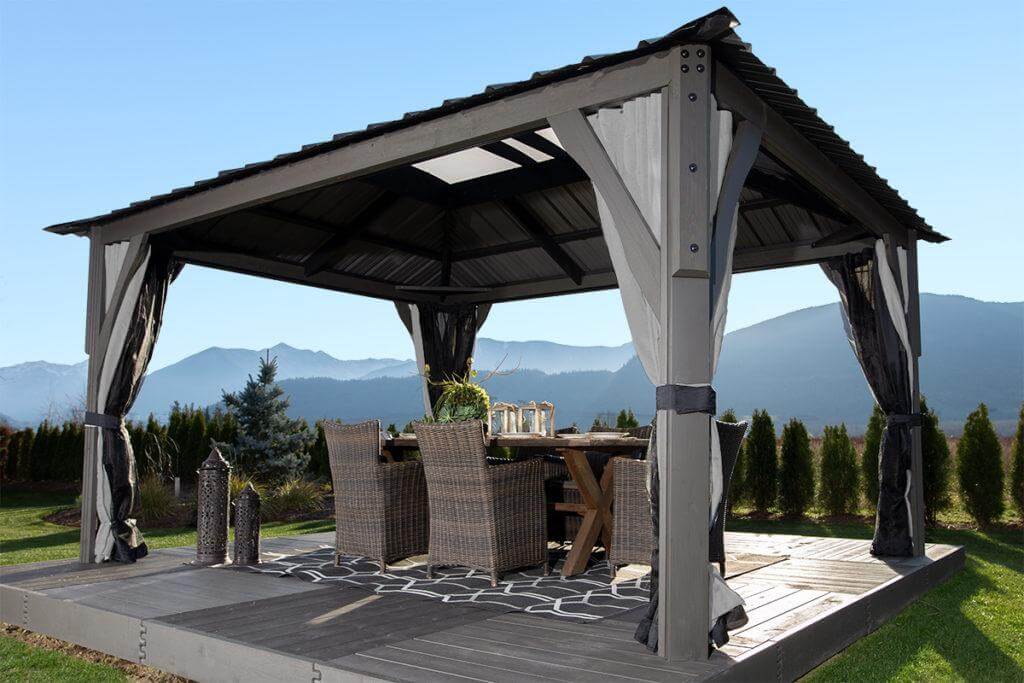 Angled side view of the Aluna gazebo, showing its square footprint with strong corner posts and sleek horizontal roof beams holding translucent roofing sheets. Inside, a modern outdoor dining set is placed centrally beneath the roof, demonstrating the gazebo’s scale and functionality for covered meals in an open garden setting.