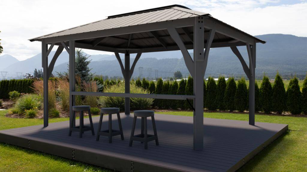 Lifestyle image of the Aluna gazebo in use, showing people seated beneath the structure around a dining table. The flat translucent roof panels provide overhead shade while leaving the setting airy and open. The clean lines of the gazebo frame the social gathering space, blending with the surrounding backyard landscaping.