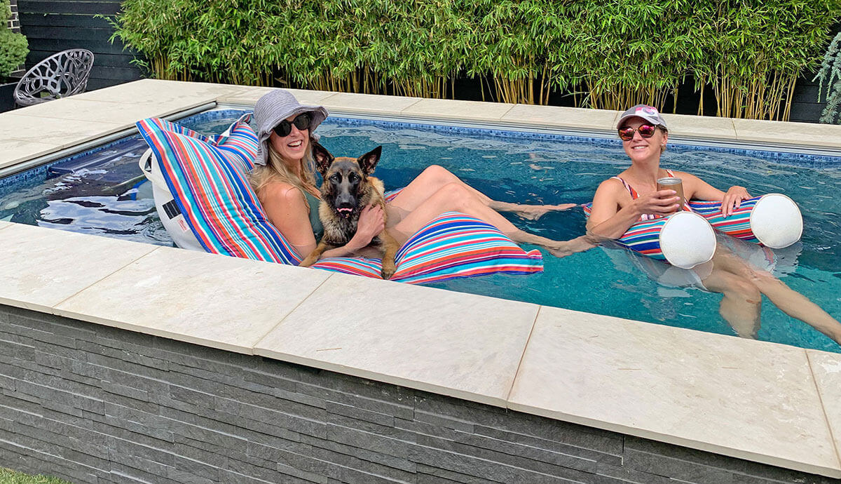 Two women lounging on colourful striped pool floats inside the Endless Pools EP207 Modular Pool, accompanied by a large German Shepherd dog. The pool is finished with grey stacked-stone exterior cladding and a cream-coloured stone coping. The bright blue water reflects a relaxing, sunny day, with tall green bamboo in the background.