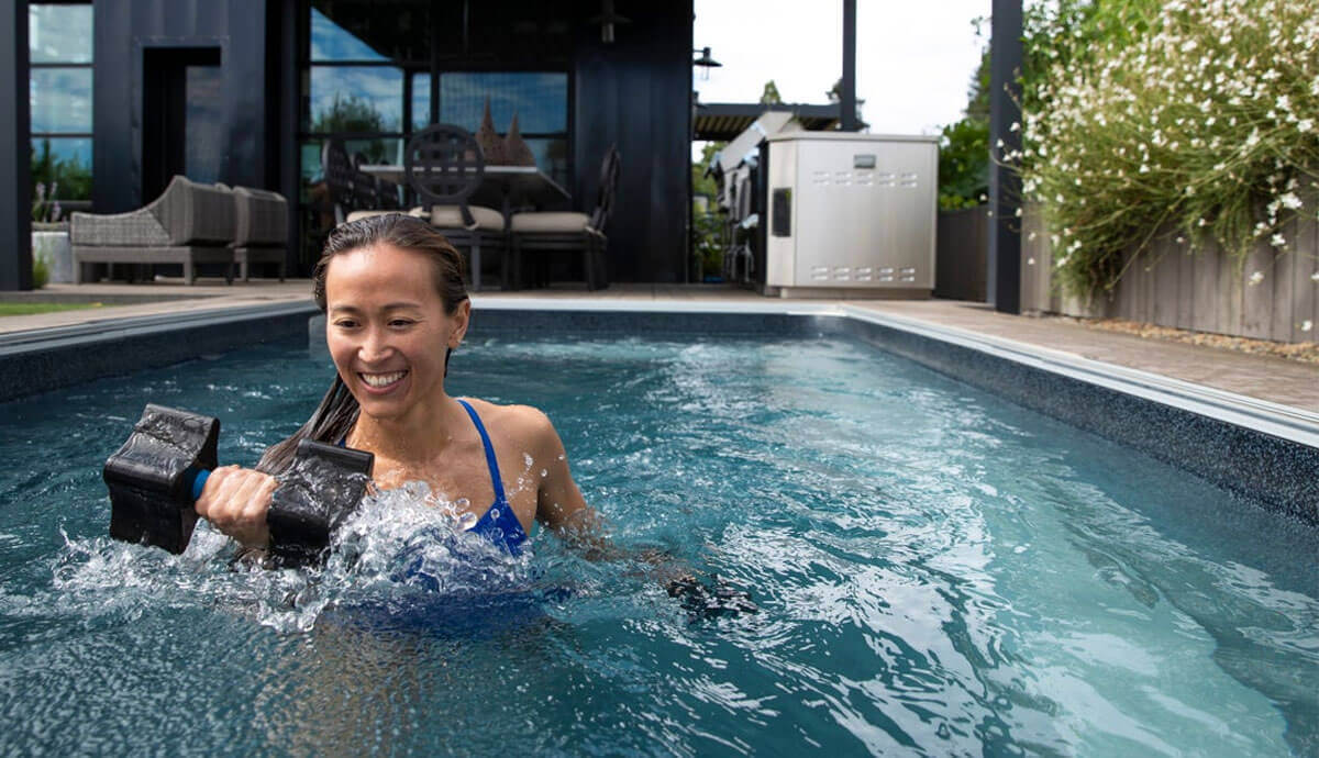 Smiling woman performing water resistance training in the EP407 Plus pool, holding two large black foam dumbbells mid-exercise. The water surface ripples around her as she moves, with the modern outdoor setting in the background featuring patio seating and lush landscaping.