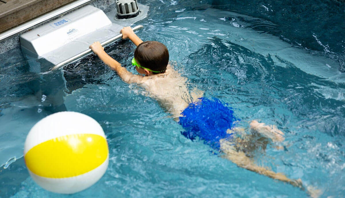 Young boy in bright blue swim shorts and green goggles practising swimming towards the Hydrodrive Plus propulsion unit. He holds the stainless-steel grab bar while water flows against him. In the foreground, a yellow-and-white beach ball floats on the clear, blue-tinted water surface.