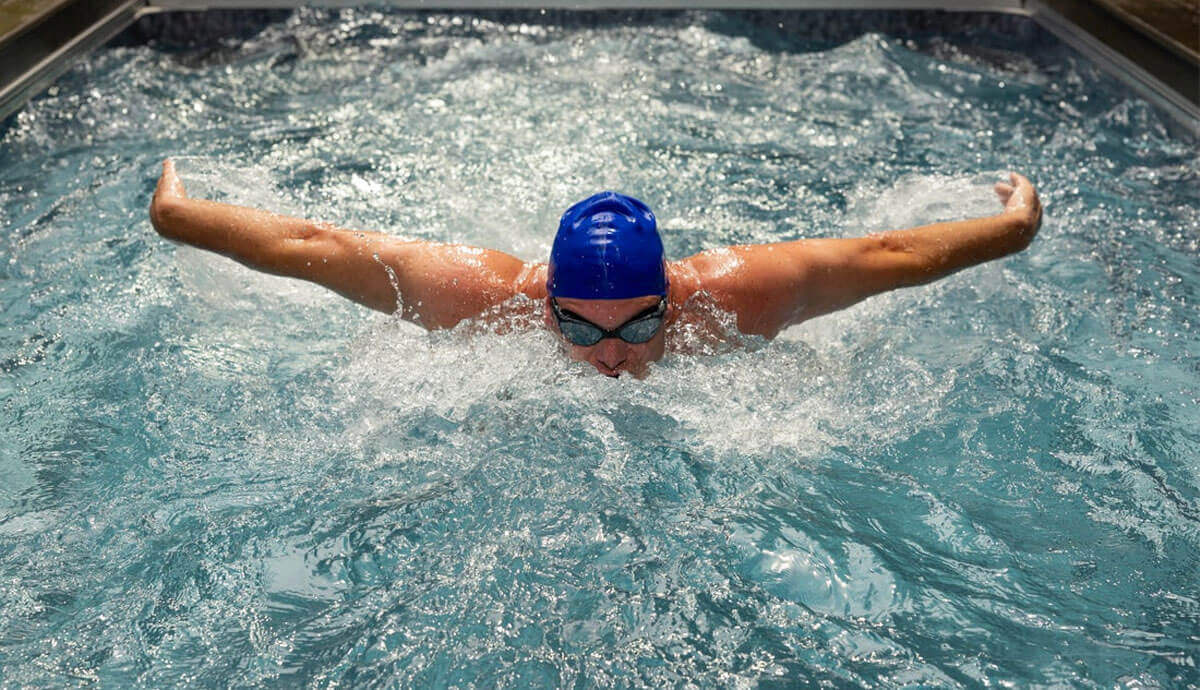 Competitive swimmer mid-butterfly stroke in the EP407 Plus pool, arms extended wide, water splashing dramatically around shoulders and chest. Wearing a blue swim cap and goggles, the swimmer moves against the adjustable current generated by the Hydrodrive Plus system.
