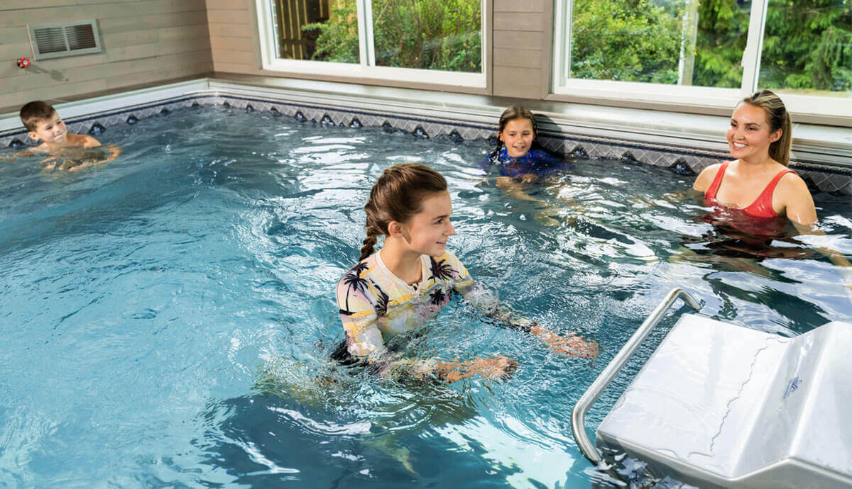 Indoor scene of a family enjoying the Endless Pools EP407 Modular Pool. The bright blue water is rippling from movement, reflecting indoor light. In the foreground, a girl wearing a tropical floral long-sleeve swim shirt stands waist-deep in the water, facing left, with both arms engaged in swimming motion. Behind her, two children and an adult woman in a red swimsuit are smiling, partially submerged, near the pool wall. The interior wall features a patterned tile border at the waterline, with windows revealing lush greenery outside. The stainless-steel propulsion system grille and handlebar are partially visible on the left side.