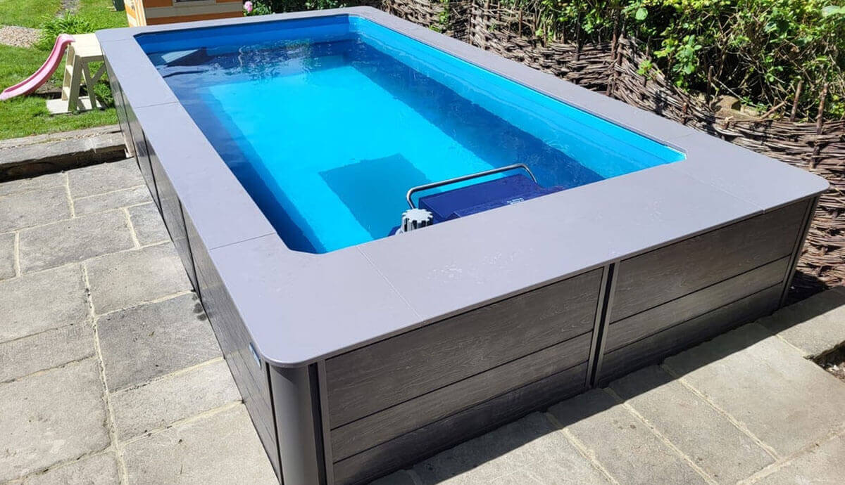 Outdoor installation of the Endless Pools EP407 Modular Pool placed on a stone patio, surrounded by a woven wooden fence and greenery. The black wood-grain composite cabinet panels are framed by silver trim, and the light grey coping contrasts with the vivid blue water inside. The Hydrodrive™ propulsion system housing is visible at the far end, with its handlebar submerged. The surrounding patio provides clear walking space, with grass and a playground slide visible in the background.