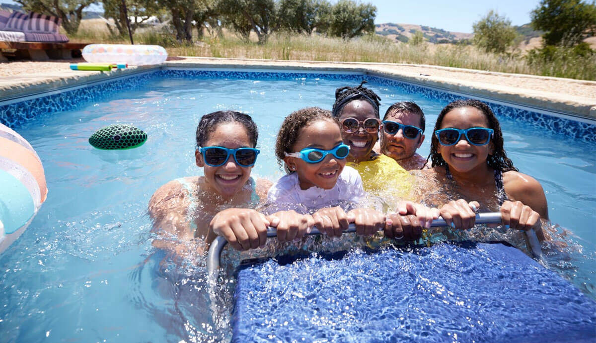 Close-up front view of five people in the Endless Pools EP407 Modular Pool, all wearing blue-tinted swimming goggles and holding onto the submerged stainless-steel handlebar attached to the propulsion system unit. The group includes two adults and three children, all smiling as water splashes around them. Bright daylight reflects off the pool’s blue interior, and floating toys are scattered in the background.
