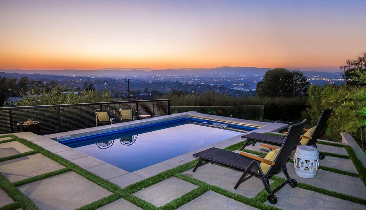 Outdoor Endless Pools EP407 Modular Pool positioned on a modern patio with grass-inlaid concrete pavers. The water reflects the orange and purple hues of the sunset over a panoramic city view in the distance. Two black mesh lounge chairs with yellow cushions are positioned at the poolside, alongside a decorative white ceramic stool. The pool’s black composite cabinet panels are trimmed with silver, and the coping frames the still, mirror-like surface of the water. A glass railing surrounds the patio, preserving the unobstructed scenic view.