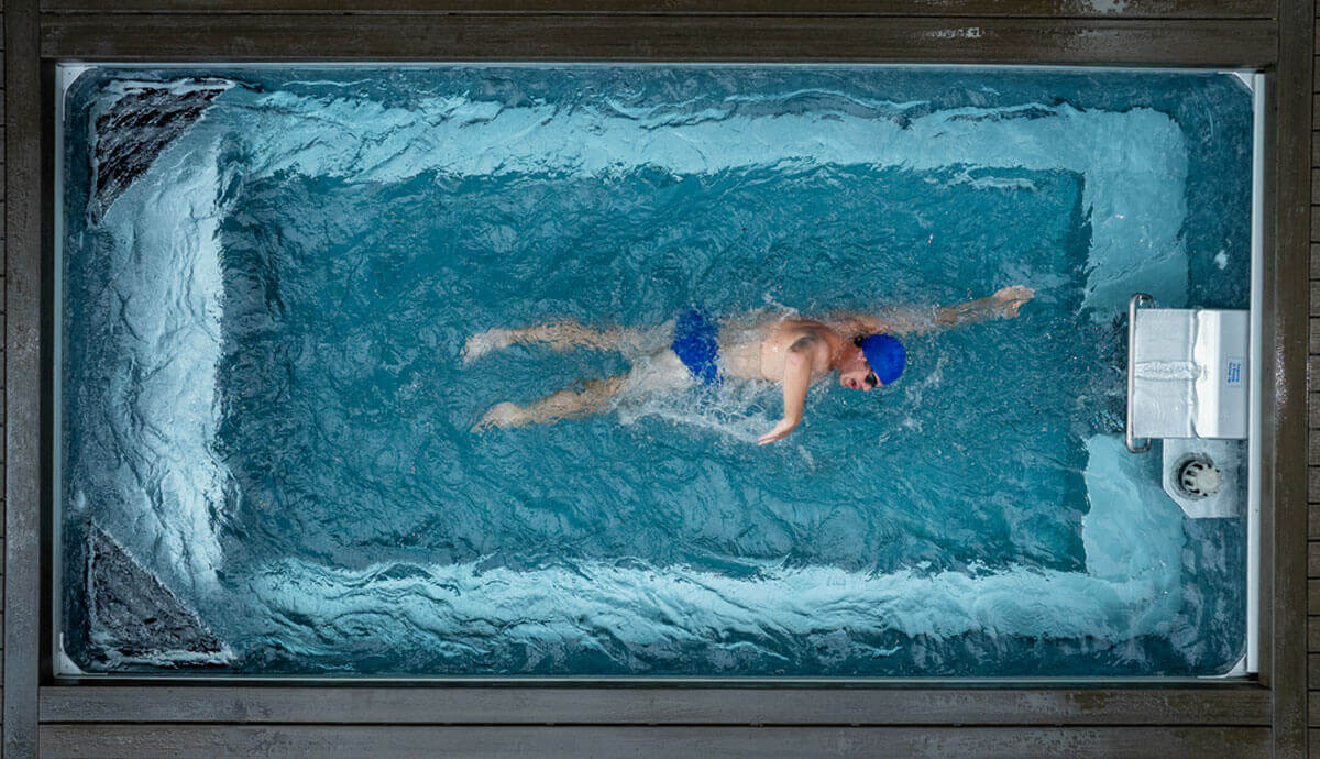 Top-down action shot of a swimmer in blue swim trunks and blue swim cap using the Endless Pools EP508 Plus Modular Pool’s Hydrodrive Plus current system. The strong, even flow creates rippling water patterns as the swimmer maintains a freestyle stroke in place.