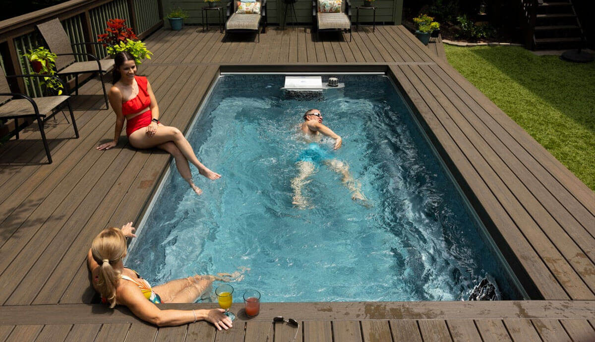 Endless Pools EP508 Plus Modular Pool installed flush within a dark brown composite deck, surrounded by outdoor lounge furniture and potted plants. Two women in swimsuits sit on the deck edge with drinks while a male swimmer in blue swim trunks uses the Hydrodrive Plus propulsion current to swim in place.