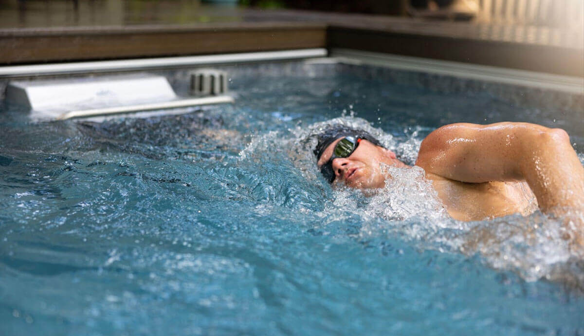 Close-up view of a male swimmer wearing mirrored goggles and a black swim cap performing a freestyle stroke against the Endless Pools EP508 Plus Modular Pool’s Hydrodrive Plus current. The propulsion grille is visible in the background, and the water surface shows dynamic splashing from the swimmer’s movement.