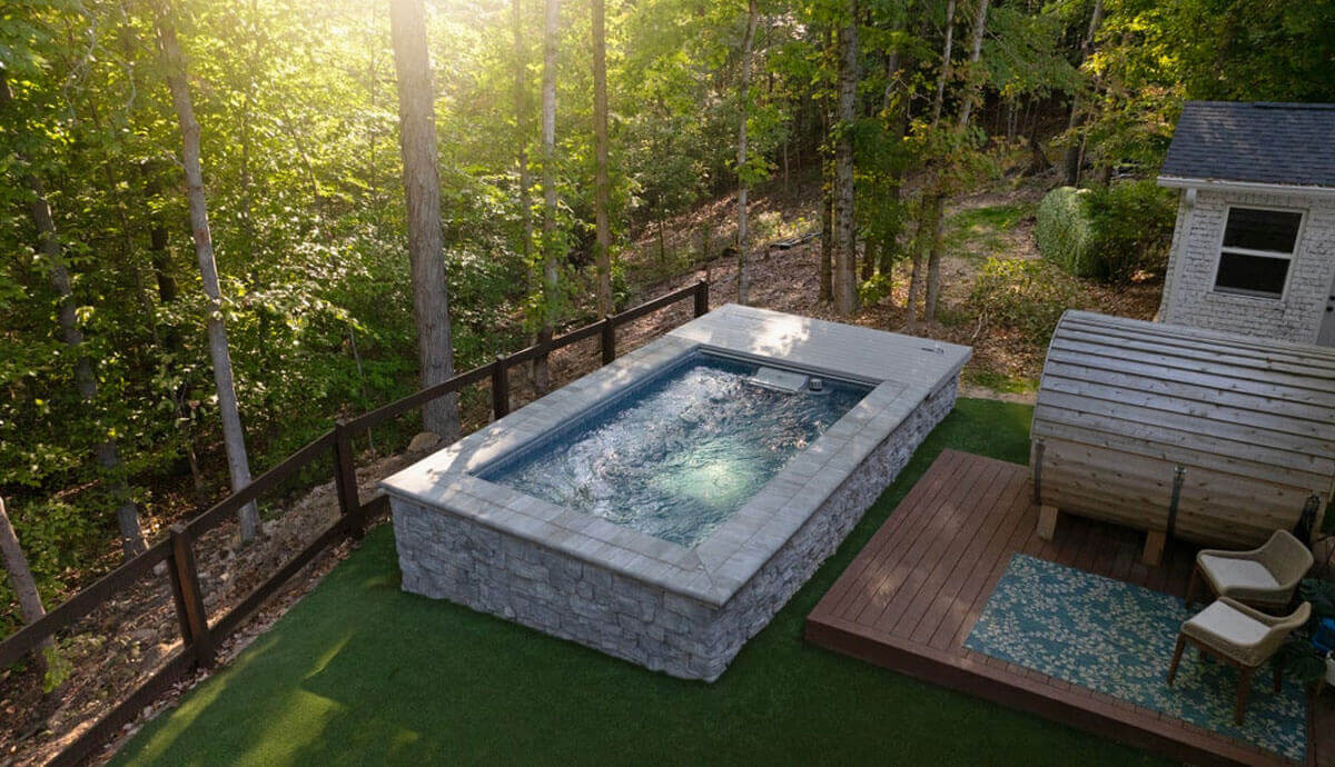Endless Pools EP508 Pro Modular Pool fully installed above-ground in a private garden setting, clad in textured light-grey stone veneer with a smooth coping edge in matching stone. The pool sits on a manicured artificial grass lawn and is bordered by a dark wooden fence on the forest-facing side. Water is active, with surface ripples caused by the operational swim current. To the right, a cedar barrel sauna stands on a raised wooden deck with a patterned outdoor rug and two armchairs. Dense trees in the background create a secluded, shaded environment with sunlight streaming from the upper left.