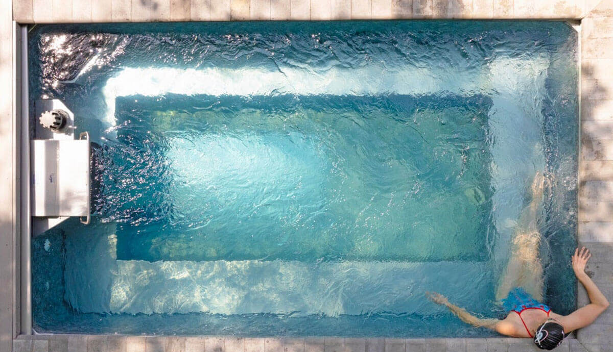 Overhead view of the Endless Pools EP508 Pro Modular Pool filled with clear, rippling water under bright natural light. At the top end, the Hydrodrive™ Pro swim current system is generating a strong, forward-facing current towards the lower end of the pool. A swimmer in a red-and-blue swimsuit is positioned at the bottom right, holding the pool edge with one hand while extending legs into the central swim lane. The rectangular layout shows the deep blue swim lane in the middle, flanked by lighter blue resting ledges and integrated steps at the bottom corners, with the coping and surrounding stone deck clearly visible.