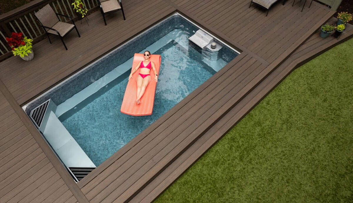 Outdoor installation of the Endless Pools EP508 modular pool, fully recessed into a brown composite deck surrounded by seating and greenery. The pool features a grey stone-look interior and is equipped with twin propulsion outlets at one end. A woman in a pink bikini reclines on an orange floating lounger, enjoying the calm, clear water under natural daylight.