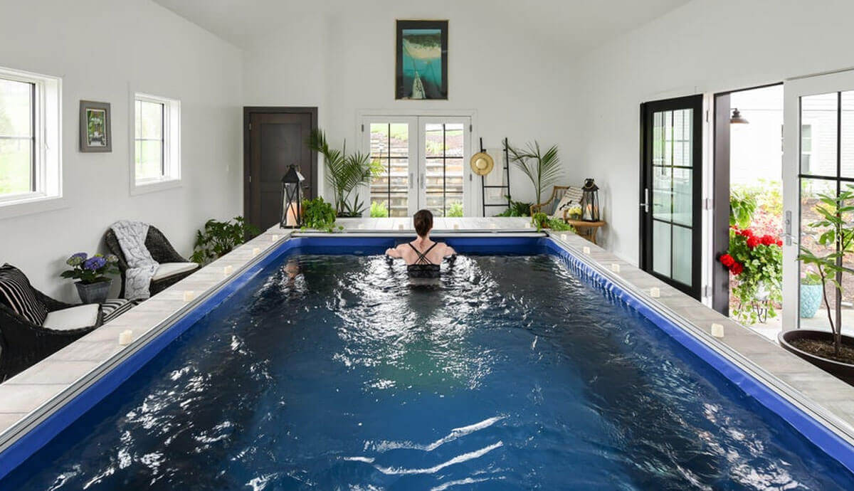 Indoor installation of the Endless Pools EP508 modular pool in a bright, modern room with white walls, multiple windows, and double glass doors opening to a garden. The pool features the vivid blue interior and Hydrodrive™ propulsion system, with a swimmer in a black swimsuit exercising in the central lane, surrounded by potted plants, wicker seating, and decorative accents.