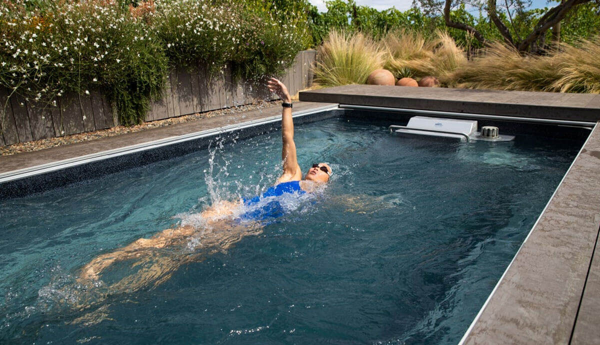 Action shot of the Endless Pools EP510 Modular Pool installed outdoors, surrounded by natural stone decking and lush greenery, with a female swimmer in a blue swimsuit performing a backstroke against the adjustable Hydrodrive™ current. Water ripples and splashes emphasise the resistance flow, while the raised current system is partially submerged at the far end.